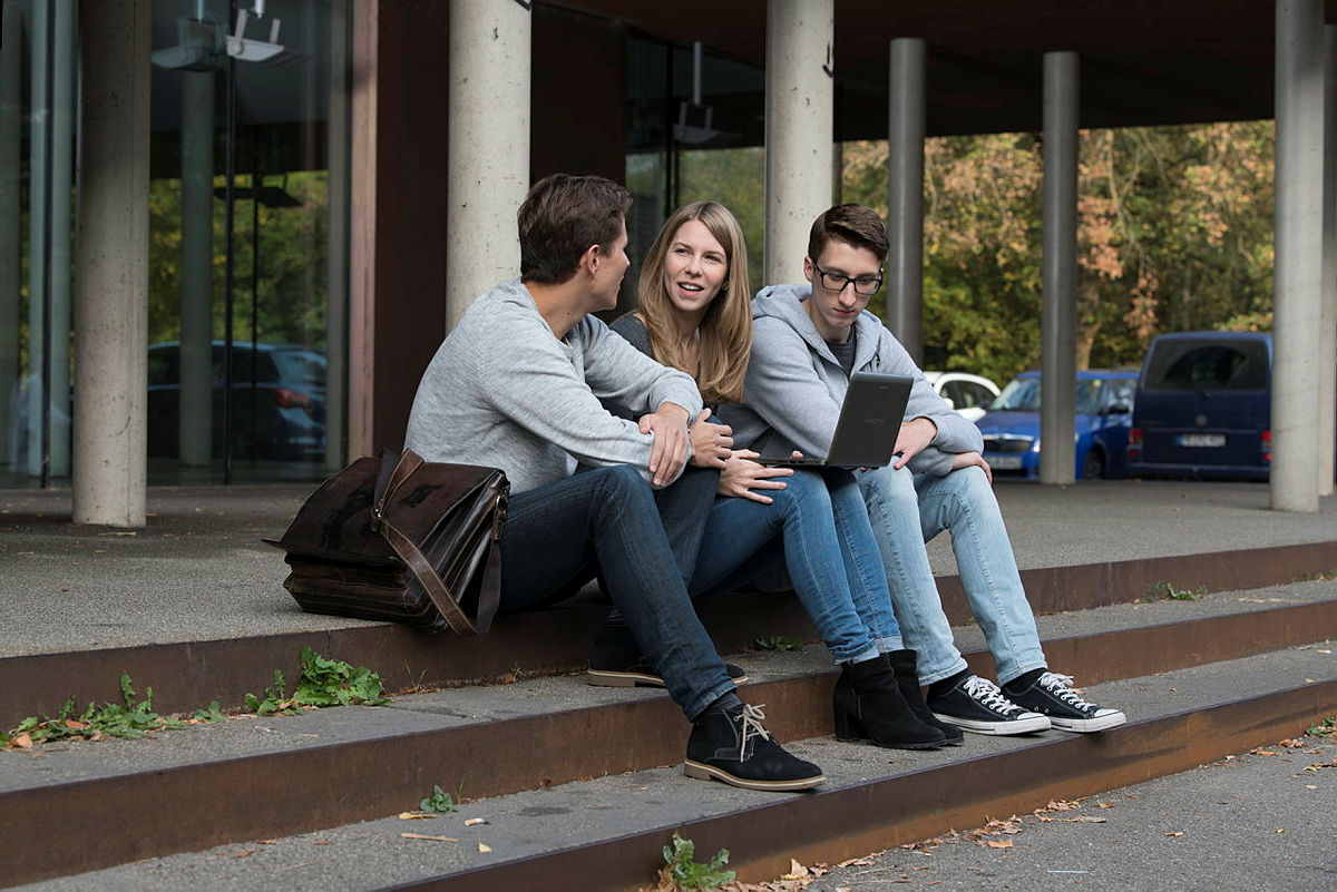 3 Studierende sitzen vor einem Uni-Gebäude auf einer Treppe